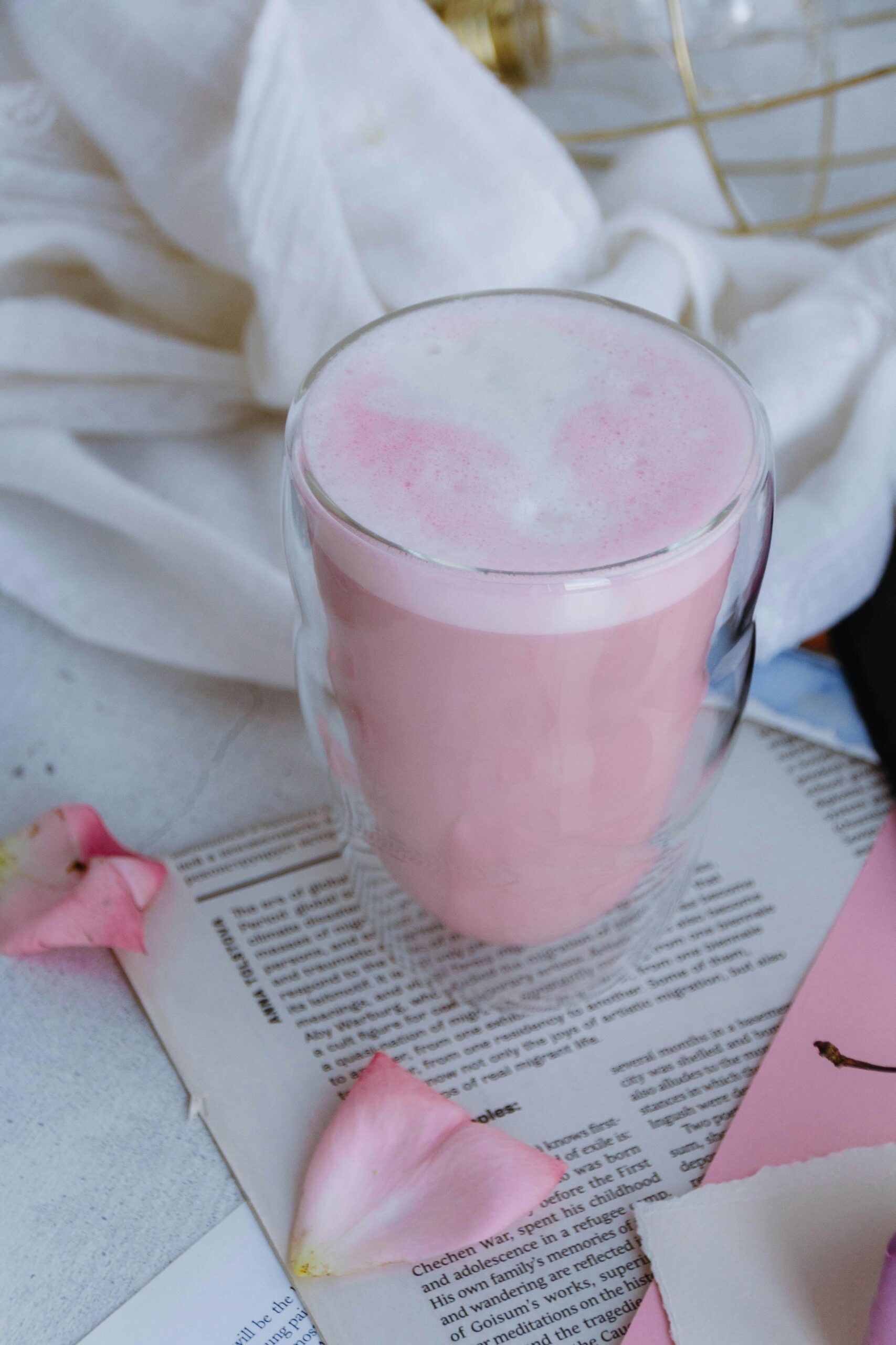 Aesthetic pink latte in a glass cup surrounded by rose petals on a cozy background.